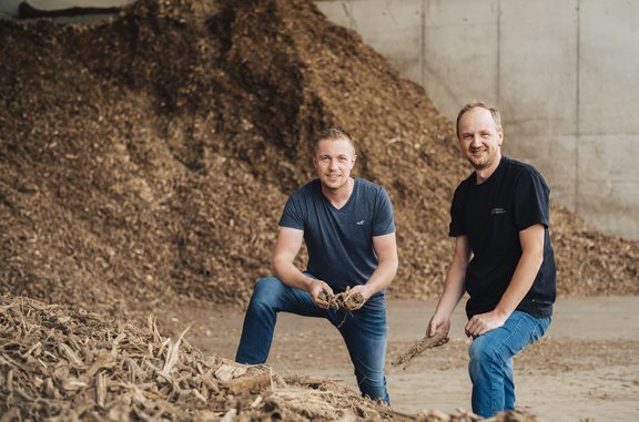 Naturwärme Zwei Männer stehen in Jeans und T-Shirt gekleidet in einer Halle in der Hackschnitzel gelagert werden. Sie lächeln in die Kamera und halten Holzhackschnitzel in der Hand.
