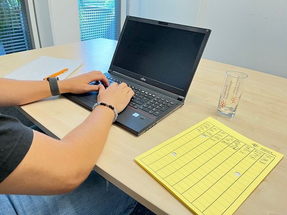 Energiesparen Person arbeitet an einem Laptop auf einem Holztisch, daneben liegt eine gelbe Mappe. Moderne Büroumgebung mit Fenster im Hintergrund.