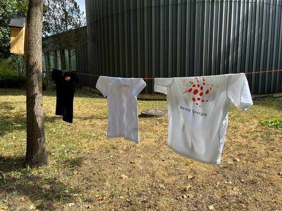 Energiesparen Drei T-Shirts hängen an einer Wäscheleine im Freien vor einem modernen Gebäude mit dunkler Metallfassade. Zwei weiße T-Shirts mit rotem Logo und dem Schriftzug 'Meine Energie.' sowie ein schwarzes T-Shirt trocknen in der Sonne. Links ist ein Baum mit einem Nistkasten zu sehen.