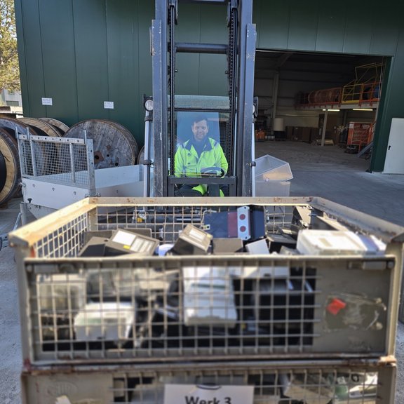 Abholung Stromzähler Ein Mann in gelber Warnjacke sitzt auf einem Stabler in einem Werkshof. Im Hintergrund sind große Kabeltrommeln zu sehen sowie eine grüne Lagerhalle. Der Stapler hat vorne auf der Gabel zwei Gitterboxen mit alten Stromzählern.
