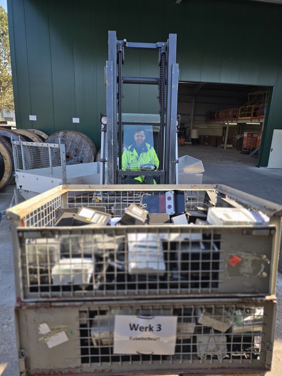 Abholung Stromzähler Ein Mann in gelber Warnjacke sitzt auf einem Stabler in einem Werkshof. Im Hintergrund sind große Kabeltrommeln zu sehen sowie eine grüne Lagerhalle. Der Stapler hat vorne auf der Gabel zwei Gitterboxen mit alten Stromzählern.