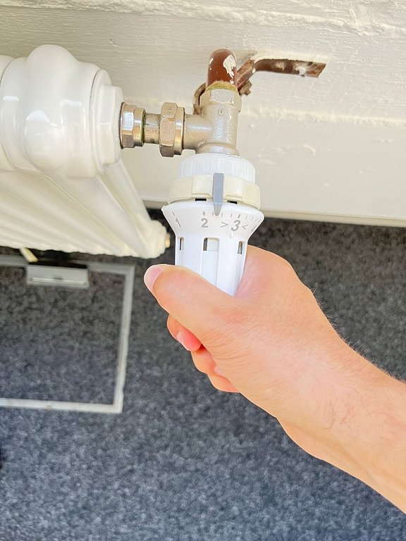 Heizung einsparen Eine Hand dreht an einem weißen Thermostat, das an einem Heizkörper befestigt ist.