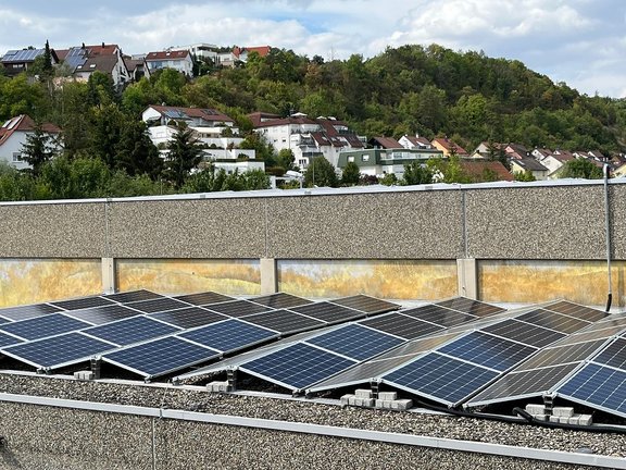 PV-Anlage Solaranlage auf einem Flachdach mit mehreren Photovoltaik-Modulen, die Sonnenenergie nutzen. Im Hintergrund eine Wohnsiedlung mit Häusern auf einem grünen Hügel. Der Himmel ist teils bewölkt.