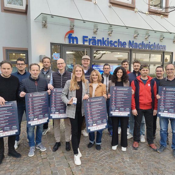 Laufgemeinschaft Tauberfranken Gruppe von Menschen steht vor dem Gebäude der „Fränkische Nachrichten“ und hält Plakate zur Volkslaufserie der Laufgemeinschaft Tauberfranken in die Kamera; Außenaufnahme auf einem gepflasterten Platz.