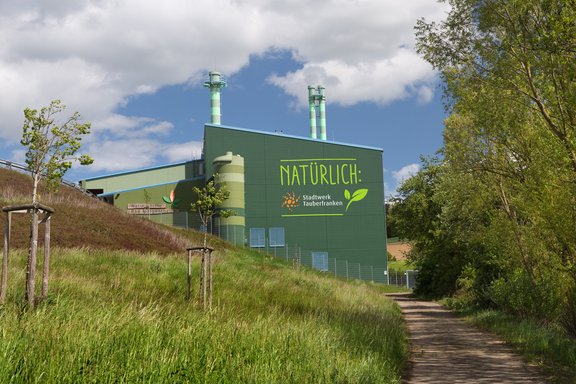 Naturwämekraftwerk Ein modernes, grünes Gebäude mit der Aufschrift „Naturwärme Bad Mergentheim“ steht unter blauem Himmel. Im Vordergrund ist eine Zufahrtsstraße und links und rechts sind satt grün Bäume zu sehen. Das Bild vermittelt den Eindruck von Nachhaltigkeit und umweltfreundlicher Energieerzeugung.