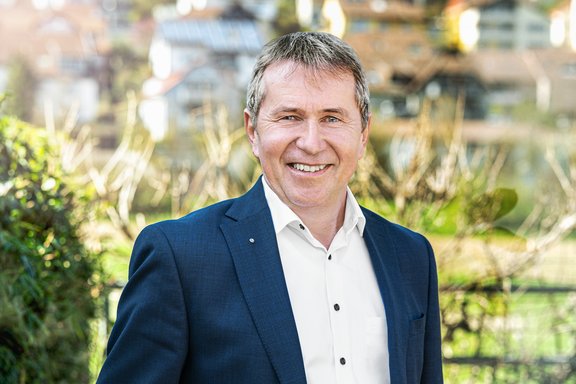 Paul Gehrig, Geschäftsführer des Stadtwerks Tauberfranken. Por Ein Mann mittleren Alters steht lächelnd mit verschränkten Armen in einem modernen, hellen Bürogebäude. Er trägt einen dunklen Anzug mit weißem Hemd und eine elegante Armbanduhr. Die Sonne scheint durch große Fenster im Hintergrund und sorgt für eine warme Lichtstimmung.