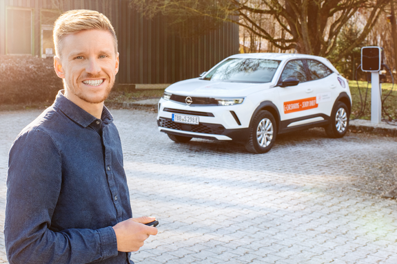 Elektromobilität Mann mit kurzem blondem Haar und blauem Hemd steht lächelnd vor einem weißen Opel SUV mit orangefarbenem Logo. Im Hintergrund ist eine Ladesäule sichtbar.